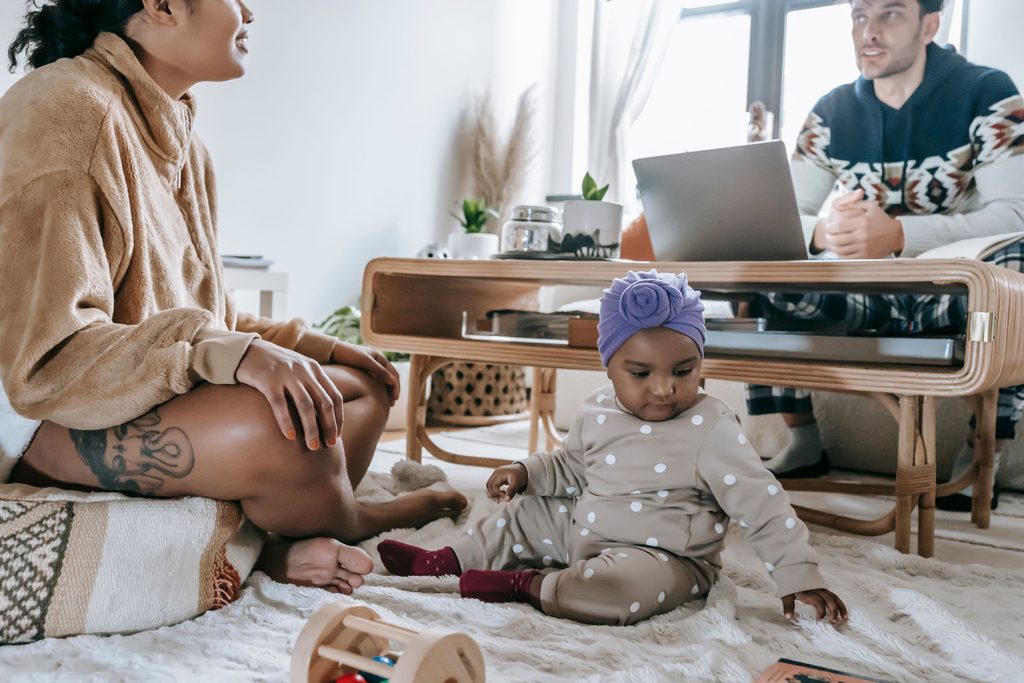 Bébé qui joue sur un tapis du salon, sous le regard de ses parents assis à côté