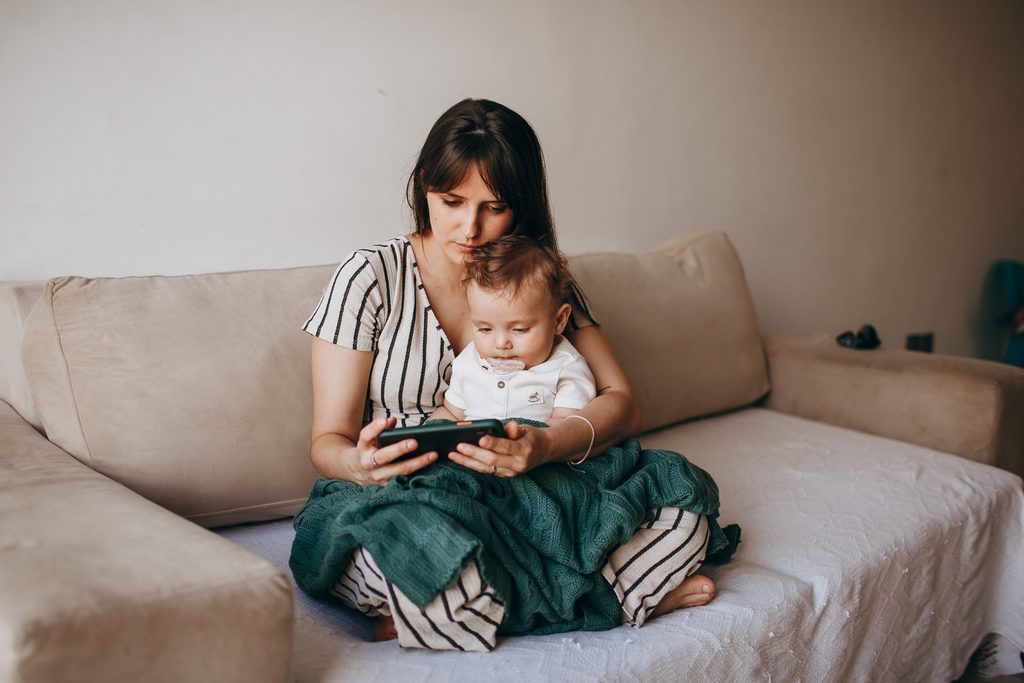Maman assise sur un canapé avec son bébé sur les genoux, en train de lire sur son téléphone